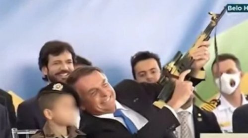 [Em cerimônia, Bolsonaro segura fuzil de brinquedo ao lado de criança vestida de militar]