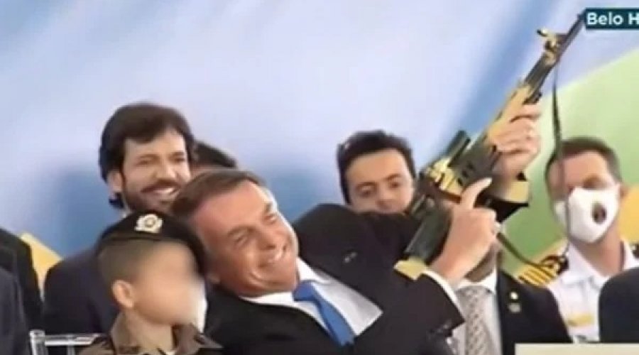 [Em cerimônia, Bolsonaro segura fuzil de brinquedo ao lado de criança vestida de militar]